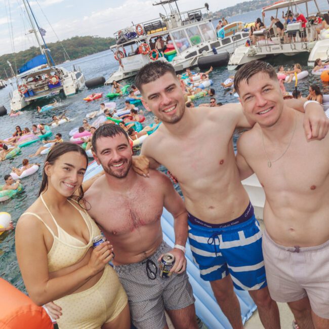 Four young adults in swimwear stand smiling on a boat at a lively floating pool party, surrounded by people on inflatables and other boats on the water under a partly cloudy sky.