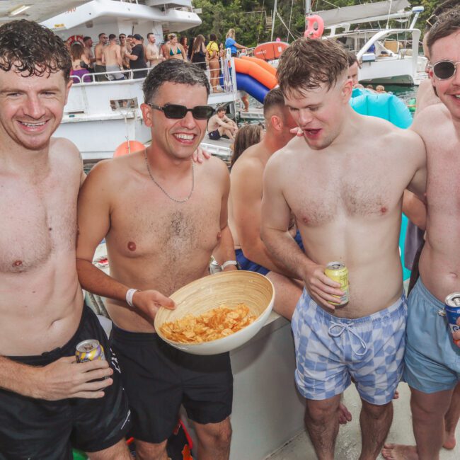 Four smiling men in swim trunks stand on a boat, holding drinks and a bowl of chips. Other people and boats are visible in the background, suggesting a lively party atmosphere on the water.
