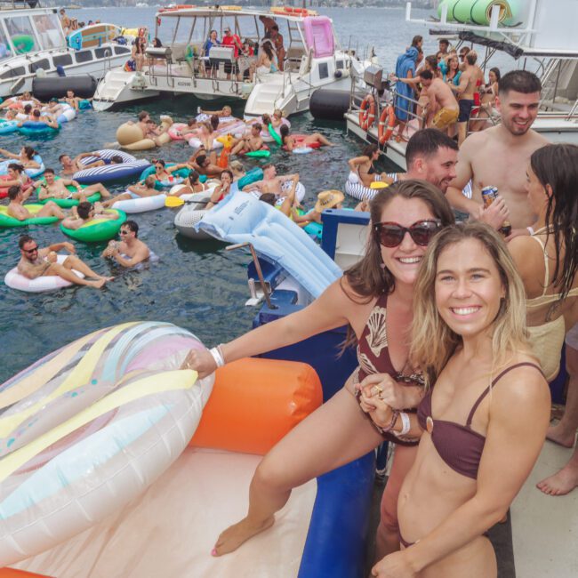 Two women in swimsuits smile at the camera on a boat surrounded by inflatable floats. Behind them, many people relax and socialize in the water and on nearby boats during a lively party.