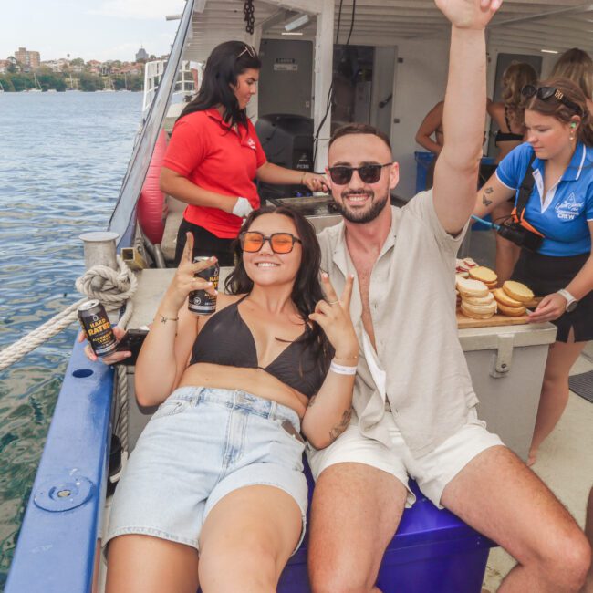 Two people sit and smile on a boat, flashing peace signs. The woman wears sunglasses, a black crop top, and denim shorts; the man wears sunglasses and light clothes. Other people and food are visible in the background near the water.