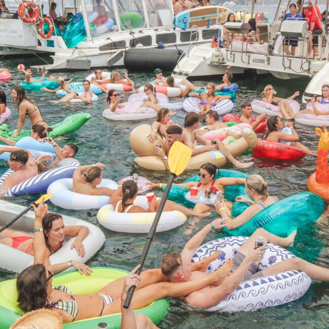 A lively group of people relax and socialize on colorful inflatable floats in the water near docked boats on a sunny day, creating a festive and vibrant summer party scene.