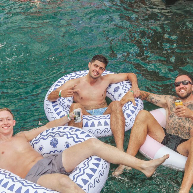 Three men relax on inflatable pool floats in clear blue water, smiling and holding canned drinks. One man flashes a peace sign, another wears sunglasses and has tattoos. A swimmer is partially visible in the background.