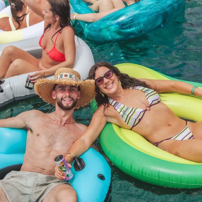 A man in a straw hat and a woman in sunglasses relax on colorful pool floats, smiling and holding drinks, surrounded by others enjoying the water.