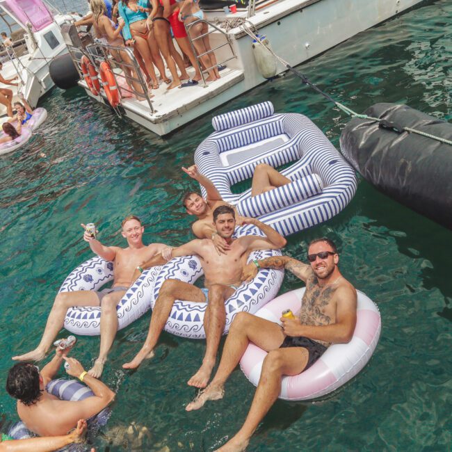 Four men relax on inflatable pool floats in the water next to a boat, smiling and holding drinks, while others socialize on the boat and nearby floats. The atmosphere is lively and festive.