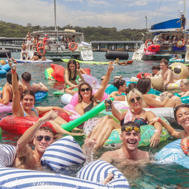 A group of people relaxing and having fun on colorful inflatable floats in the water, holding drinks and smiling, with boats and trees visible in the background on a sunny day.