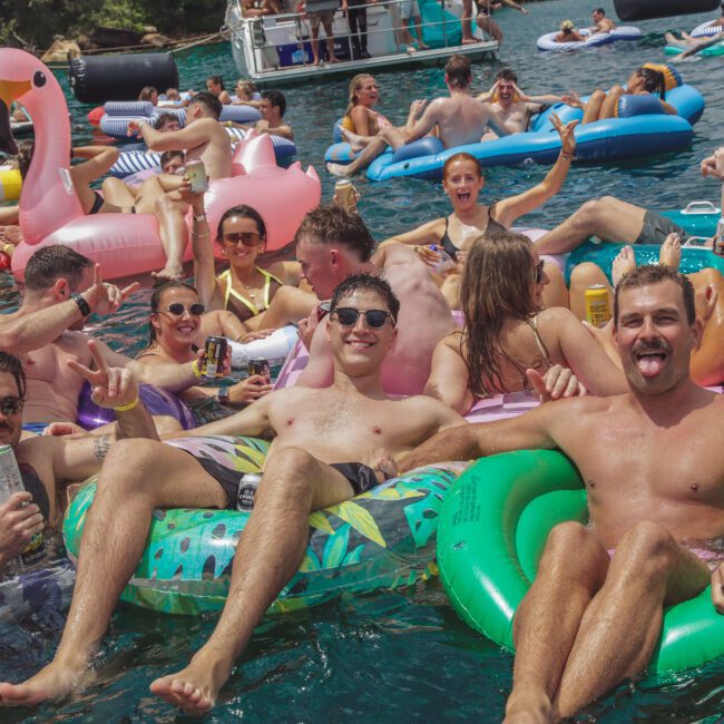 A large group of young adults relax and have fun on colorful inflatable floats in bright blue water, smiling, laughing, and holding drinks at a lively summer pool or boat party.