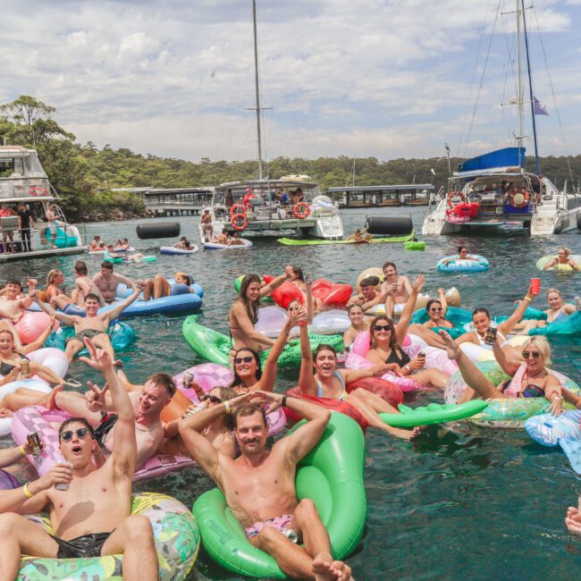 A large group of people on colorful pool floats enjoying a party in the water, smiling and raising drinks, with boats and a dock in the background surrounded by trees under a partly cloudy sky.