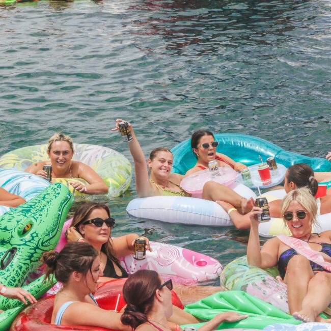 A group of women relax on colorful pool floats, smiling and raising drinks in celebration. The water is clear, and the scene appears festive, with many floats shaped like animals and a cheerful, party atmosphere.