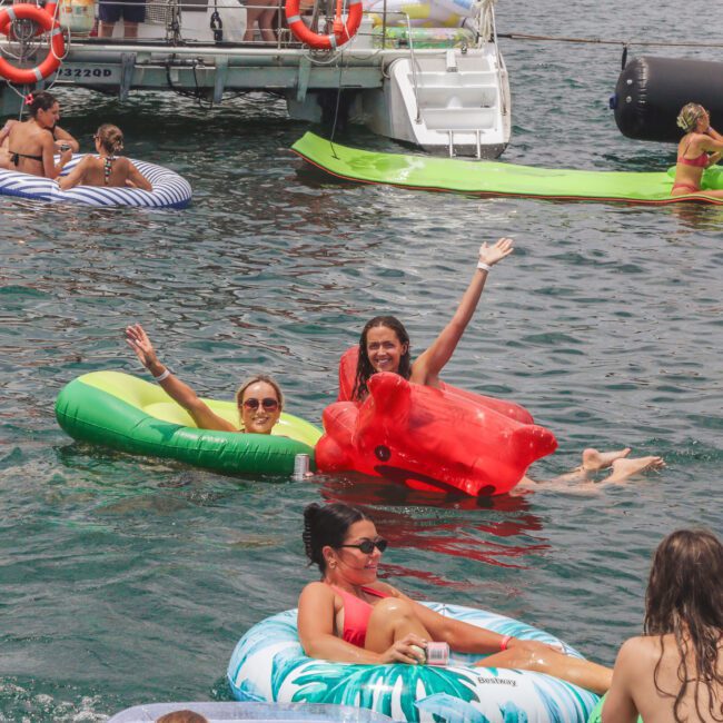 People relaxing on colorful inflatables in the water near a boat. Three women smile and wave at the camera, while others lounge and swim nearby under a sunny sky.