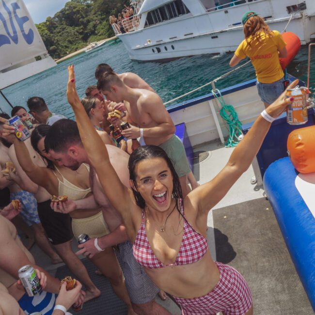 A woman in a red checkered bikini smiles and raises her arm on a crowded party boat. People around her eat, drink, and socialize, with a pool float and ocean in the background. A yacht is visible nearby.