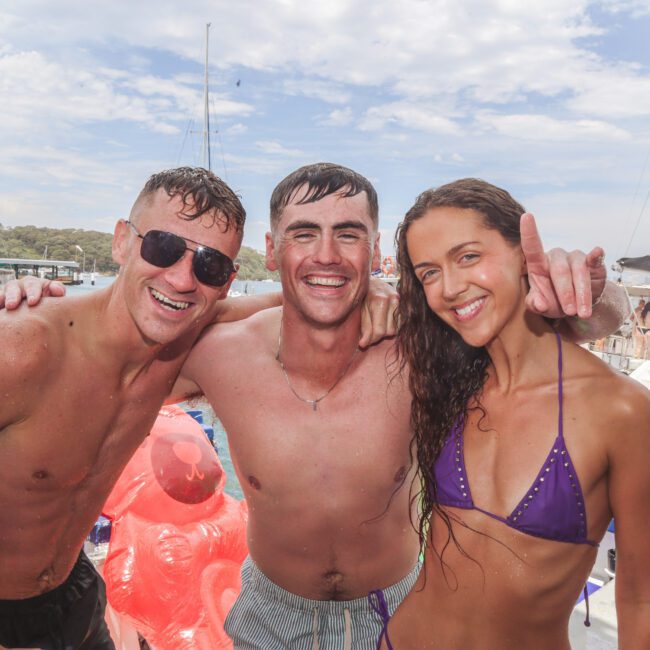 Three young adults in swimwear smile at the camera on a boat, with one woman holding up her index finger. Other people, boats, and water floaties are visible in the background on a sunny day.