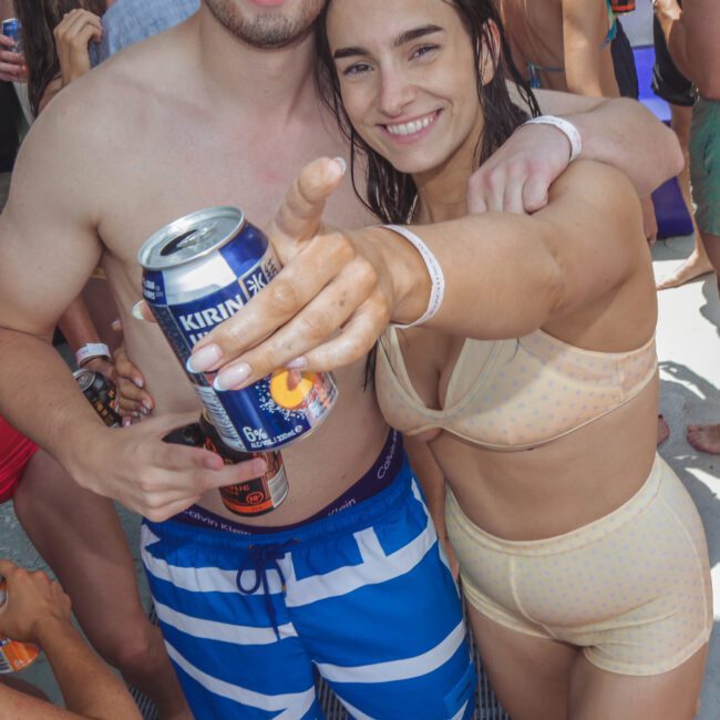 A man in blue striped swim trunks and sunglasses stands with a smiling woman in a light-colored bikini, both holding drinks and posing for a photo at a crowded beach or pool party.