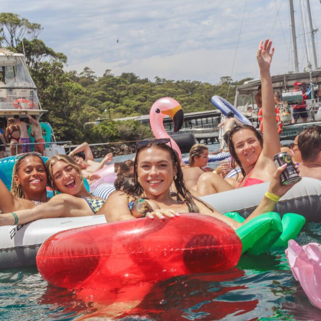 A group of smiling young people relax on colorful pool floats in the water, holding drinks, with boats and lush greenery in the background on a sunny day.