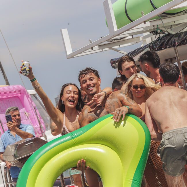 A group of people at a lively boat party, smiling and holding drinks and a green inflatable float, with others relaxing and socializing in the background under a sunny sky.