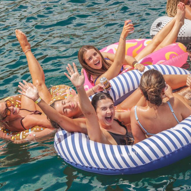 A group of smiling young adults relax on colorful inflatable pool floats in clear blue water, waving and holding drinks as they enjoy a sunny day together.