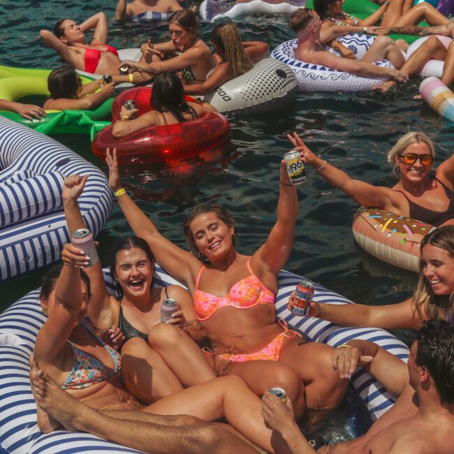 A group of people relax and smile on colorful inflatable pool floats in a body of water, holding drinks and raising their arms in celebration on a sunny day.
