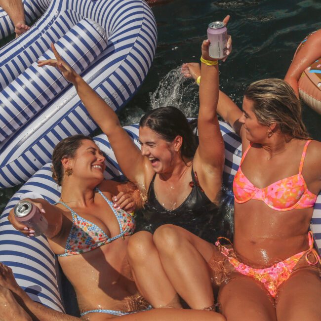 Three women in swimsuits relax on inflatable pool floats, smiling and holding drinks, surrounded by others enjoying a sunny day in the water.