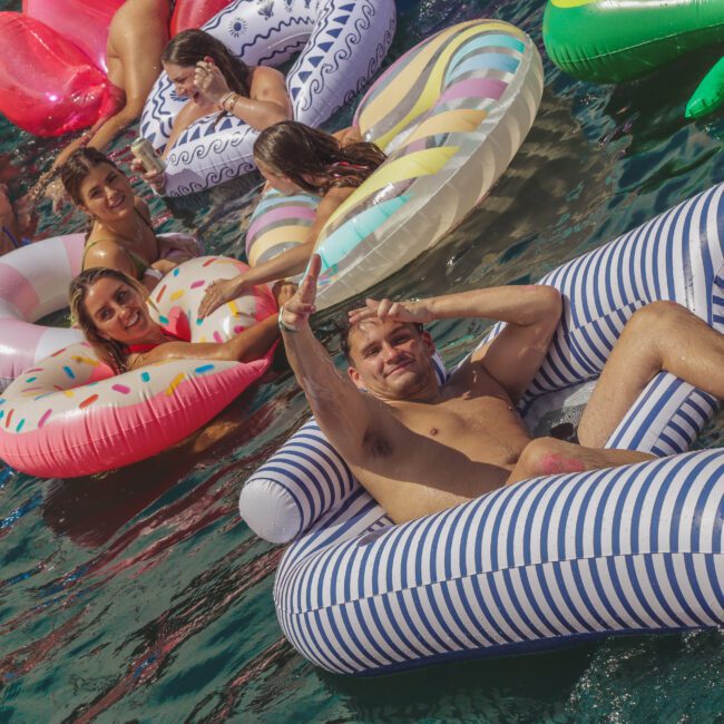 People relax on colorful inflatable pool floats in the water. One man in the foreground smiles and makes a peace sign, while others lounge and enjoy the sunny day. The atmosphere is lively and fun.