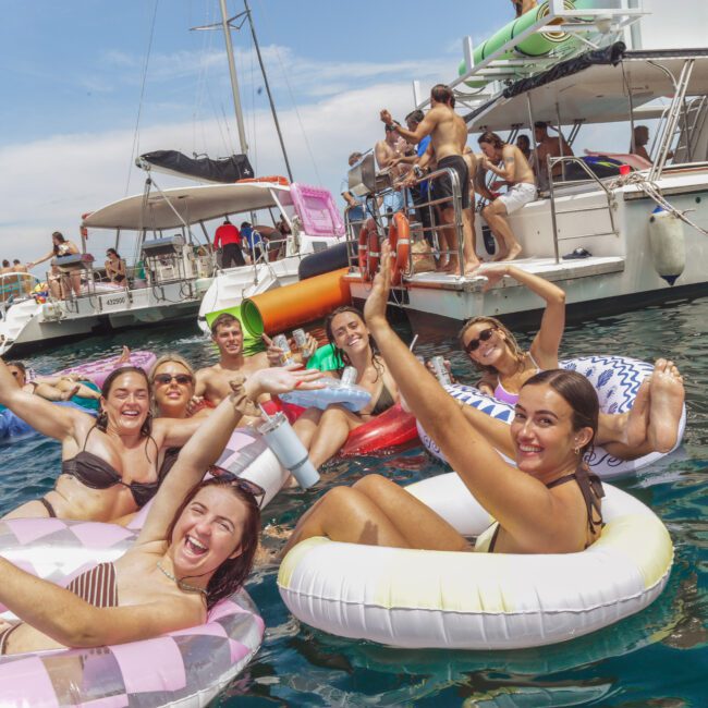 A group of smiling people relax on colorful floaties in the water, waving at the camera, with boats and more people in the background enjoying a lively party under a sunny sky.
