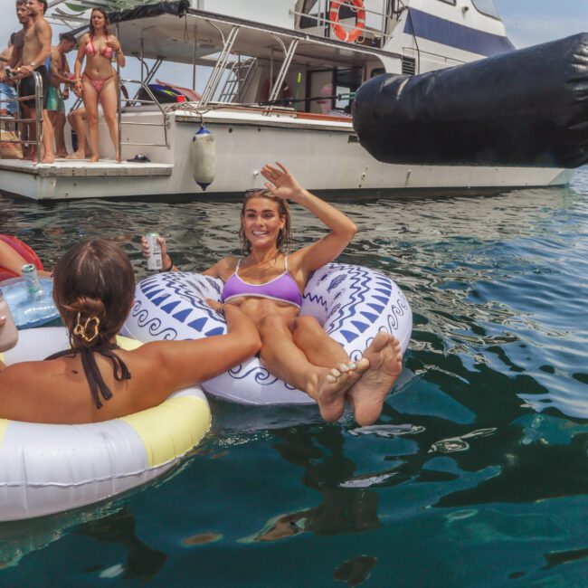A woman in a bikini smiles and waves while floating on an inflatable tube in the water near a boat. Other people are on inflatables and standing on the boat in the background, enjoying a sunny day.