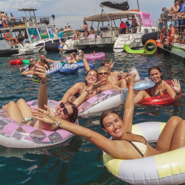 A group of people in swimsuits float on colorful inflatable rings in the water, smiling and waving at the camera. Boats with more people are anchored nearby under a partly cloudy sky.