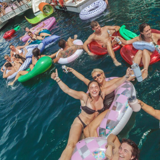 People relaxing on colorful pool floats in the water near boats. Many are smiling, wearing swimsuits, and holding drinks, enjoying a sunny day. The scene is lively and festive with a mix of men and women.