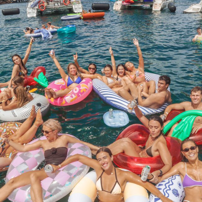 A group of young adults relax and pose on colorful inflatable floats in clear blue water, surrounded by boats. Many are smiling, holding drinks, and wearing swimwear, enjoying a sunny day outdoors.
