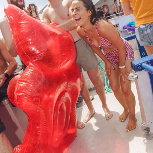 A group of young adults laughing and enjoying themselves at a pool party on a boat, holding a large red inflatable float. One woman is wearing a red-checkered swimsuit and a lifeguard stands in the background.