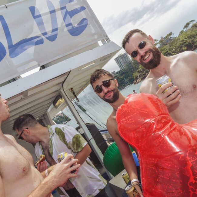 Four men are socializing on a sunny boat party. Two are smiling at the camera, holding drinks, one has a red inflatable float, and a "BLUE" banner is visible above them. Water and greenery are in the background.