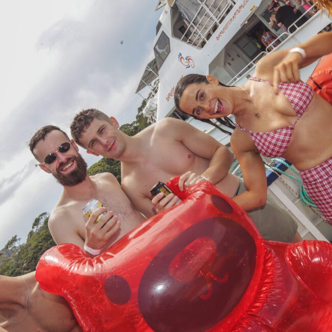 Three young adults in swimwear smile and pose with drinks on a boat deck. A woman in a red-checkered bikini is holding a large red inflatable bear. A white yacht and trees are visible in the background.