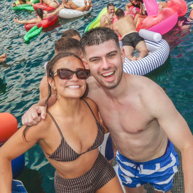 A smiling woman in sunglasses and a striped swimsuit stands beside a smiling man in blue striped swim trunks on a boat ladder, with people relaxing on colorful inflatables in the water behind them.