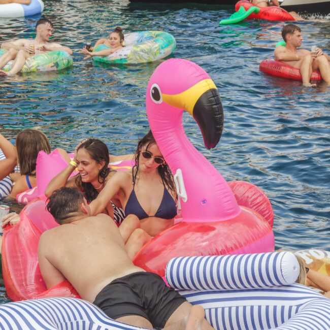 People relax on colorful inflatable floats, including a large pink flamingo, in a pool. Some are talking, laughing, or lounging in swimwear, enjoying a lively summer gathering on the water.