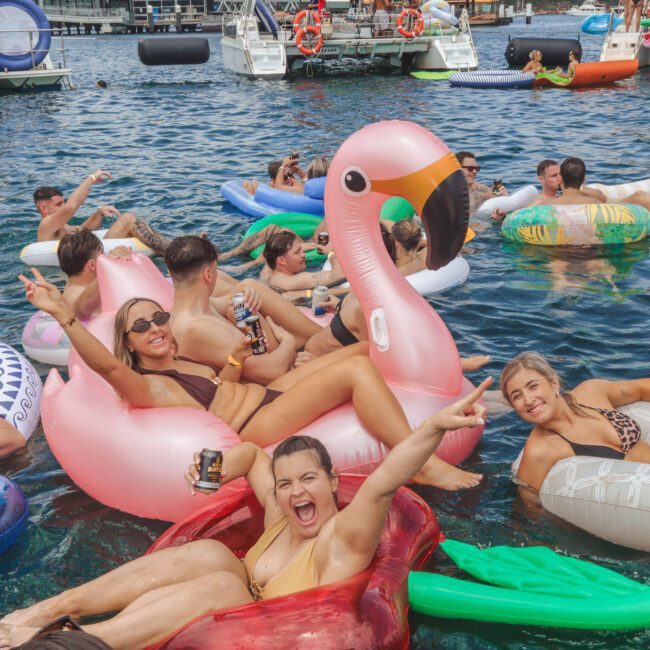 A group of people in swimsuits relax and celebrate on colorful inflatable pool floats in the water, including a pink flamingo and a red chair, with boats and more floats in the background. Everyone appears happy and festive.