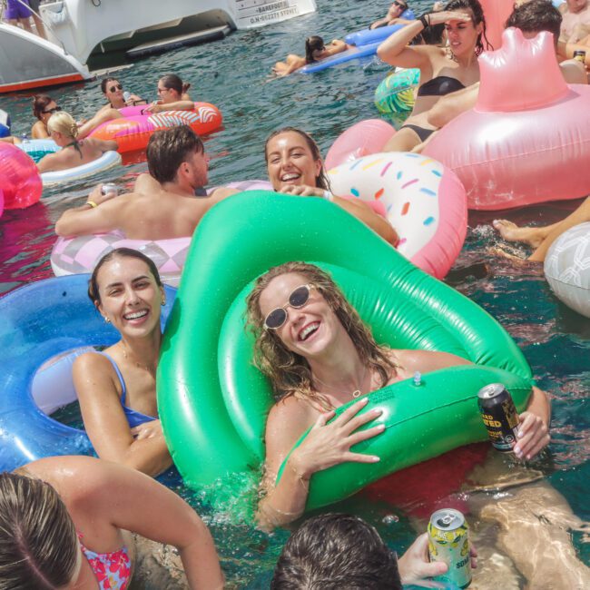 People enjoying a pool party on the water, surrounded by colorful inflatable floats shaped like donuts, flamingos, and rings. They are smiling, laughing, and holding canned drinks under the sun.