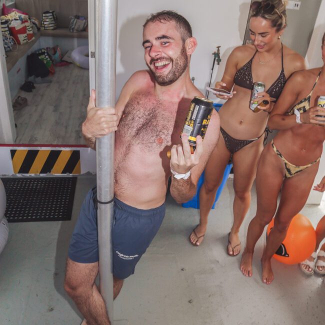 A smiling man in swim trunks poses playfully with a can, holding onto a pole on a boat. Behind him, people in swimsuits laugh and hold drinks. The setting appears festive and relaxed.