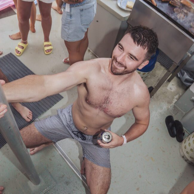 A man in swim trunks holds a drink and smiles while climbing a metal ladder on a boat. Around him, several people in swimsuits and casual wear are standing and talking. Grilled food and drinks are visible in the background.