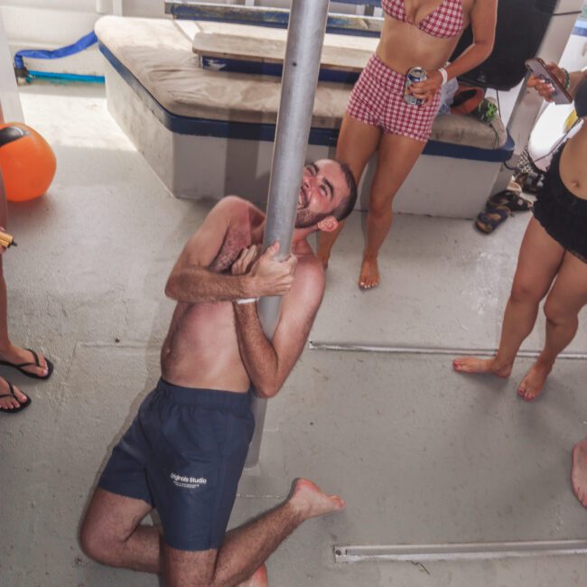 A man in swim trunks playfully wraps around a metal pole on a boat deck while laughing. Several people in swimsuits stand nearby, smiling and watching. The setting appears festive and relaxed.