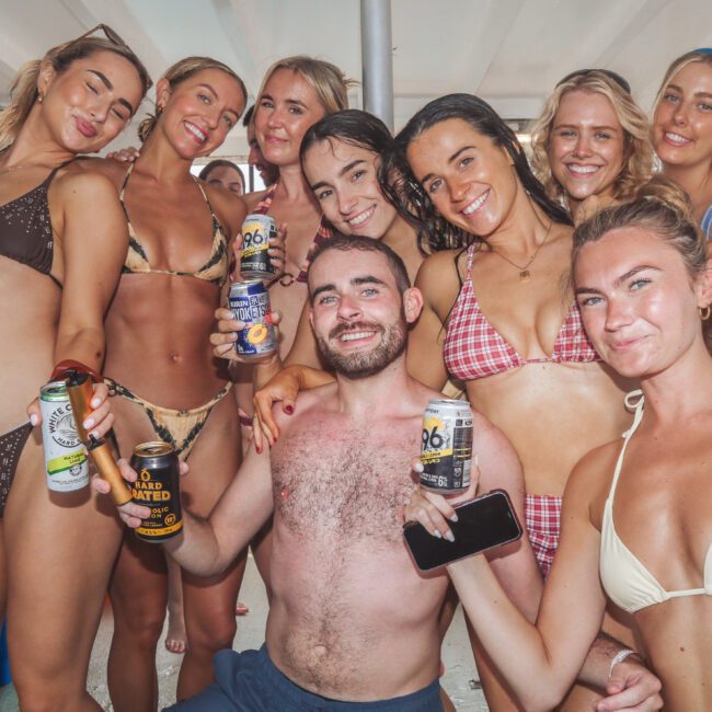 A group of smiling young adults in swimsuits pose together on a boat, holding drinks and enjoying a sunny day. The group appears happy and relaxed, suggesting a fun social gathering or vacation.