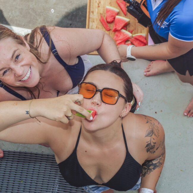 Two women in swimwear smile up at the camera; one is eating a slice of watermelon. Others stand nearby, and there are pieces of watermelon on a wooden board in the background.