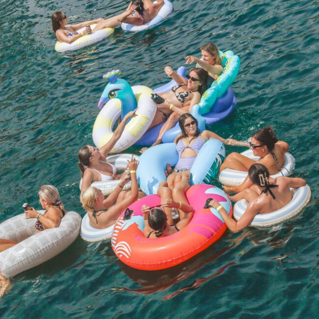 A group of people relax on colorful inflatable pool floats in a body of water, enjoying drinks and sunshine. The scene is lively, with various float shapes and friends socializing in a summer setting.