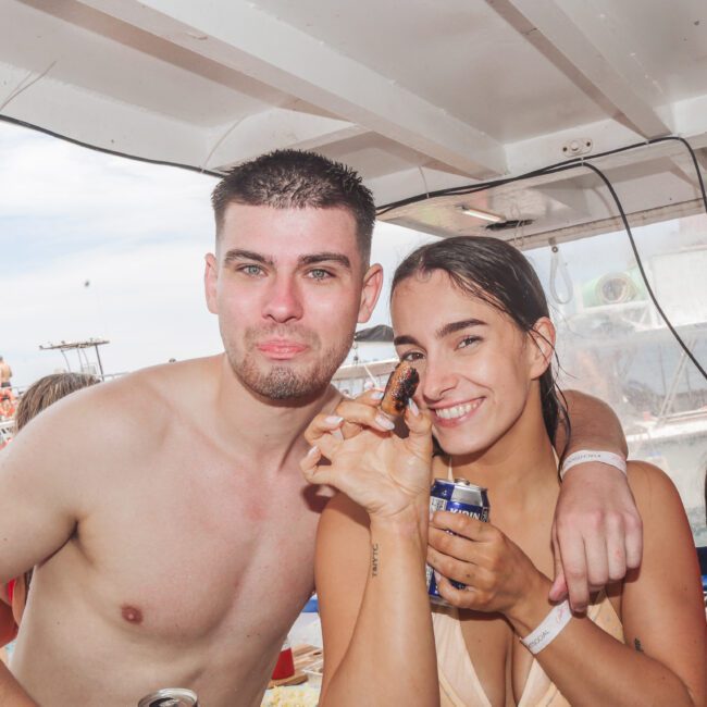 A smiling woman in a swimsuit holds a drink and food, standing next to a shirtless man on a boat. Other people and boats are visible in the background, with blue skies overhead.