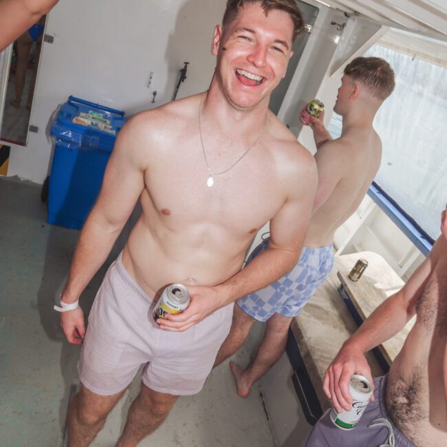 A smiling, shirtless man in pink swim trunks holds a can and stands on a boat with other people nearby, all appearing relaxed and enjoying a sunny day.