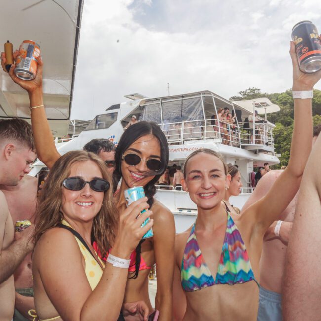 A group of people in swimwear smile and raise drinks on a crowded boat party, with another boat and greenery visible in the background. Everyone appears to be enjoying a sunny day on the water.