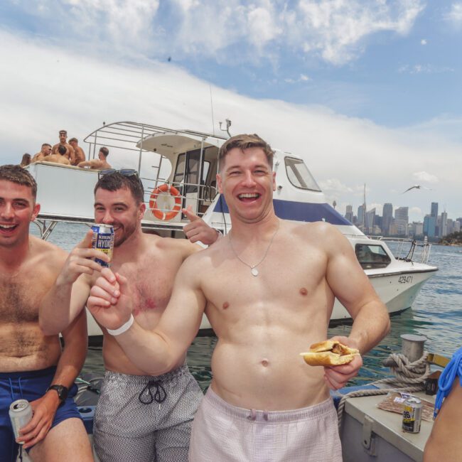 Four people in swimsuits smile and laugh on a boat, holding drinks and food. Other boats and people are visible in the background on the water, with a city skyline and partly cloudy sky behind them.