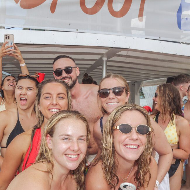 A group of young adults in swimsuits smile and pose for a photo on a boat, some holding drinks. One person is taking a selfie while others laugh and enjoy the sunny atmosphere.