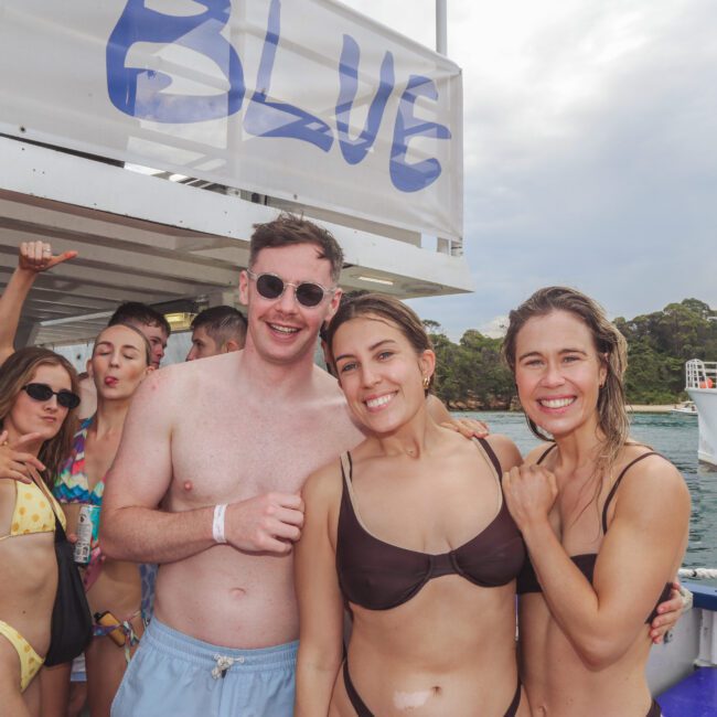 A group of young adults in swimsuits smile and pose together on a boat, with the ocean and another boat in the background. The atmosphere is lively and sunny, suggesting a fun outing or party.