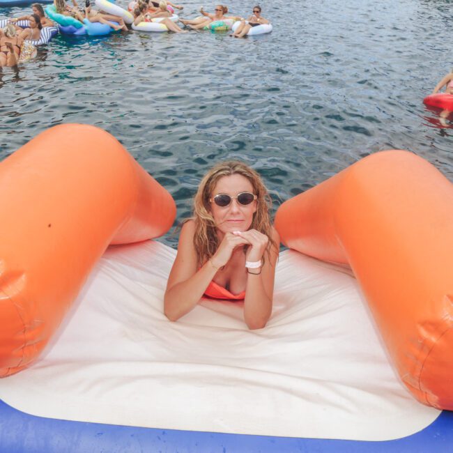 A woman with curly hair and sunglasses relaxes on a large orange float in the water. Other people on colorful inflatables are in the background, enjoying a sunny day.