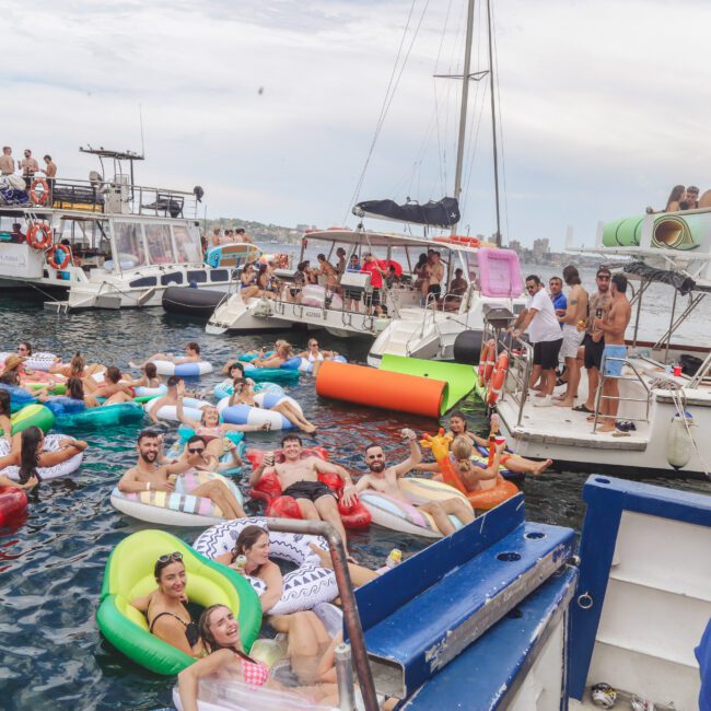 A lively group of people relax on colorful pool floats in the water near several anchored boats, enjoying a festive atmosphere on a partly cloudy day. Some people are on the boats, while others lounge and chat in the water.