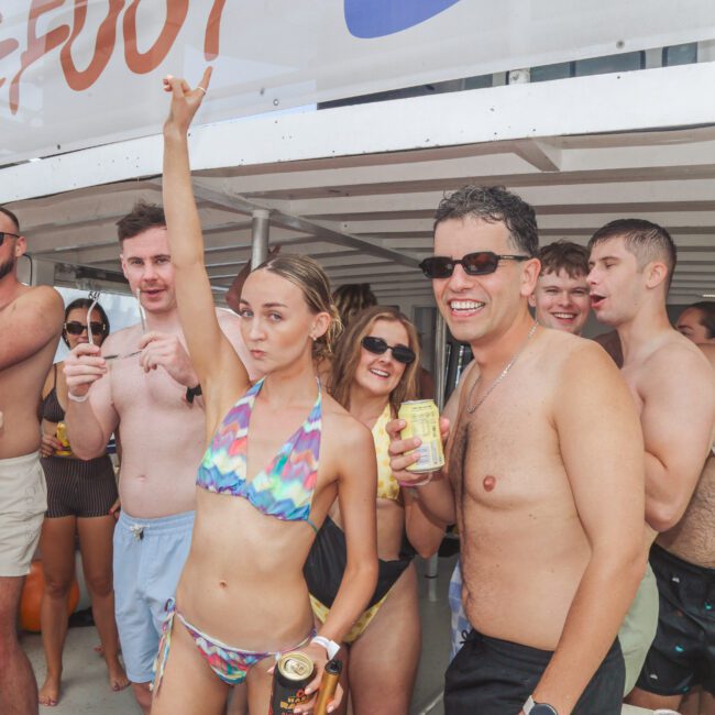 A group of people in swimsuits smile and pose together at an outdoor party, holding drinks. One woman raises her arm in the air. The atmosphere is lively and festive under a white canopy.