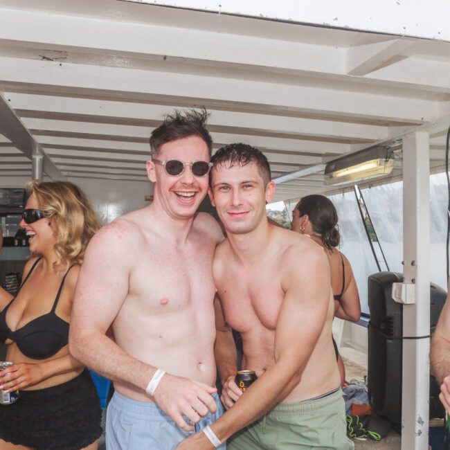Two young men, shirtless and smiling, stand close together on a boat surrounded by other people in swimwear, some holding drinks, enjoying a lively and sunny party atmosphere.